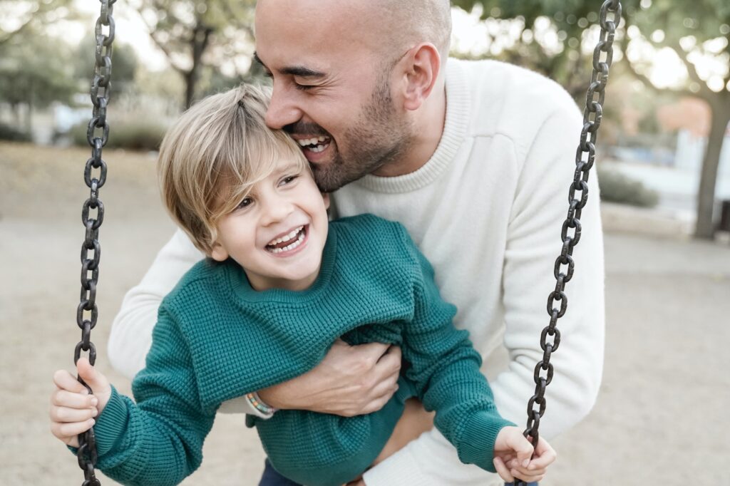 Happy dad and son having fun in city park swinging on swing - Main focus on father eye